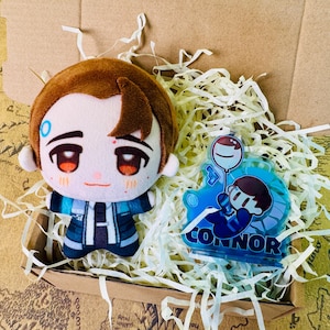 May include: A plush doll and a blue acrylic charm featuring a character design. The doll has brown hair and a blue and white outfit. The charm has the name "CONNOR" and a cartoon illustration. Both items are in a cardboard box with paper filler.