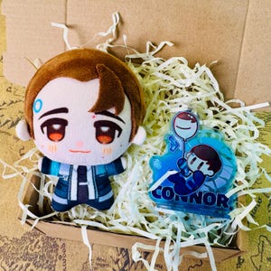 May include: A plush doll and an acrylic charm featuring a character with brown hair and blue accents. The doll is in a cardboard box with paper filler. The charm has the name "CONNOR" on it.