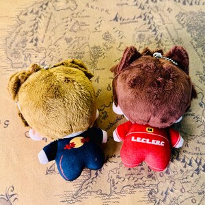 Peut inclure: Deux poup&eacute;es en peluche avec des cheveux bruns et beiges, vues de dos. L'une porte une tenue bleu marine avec un motif rouge et jaune, l'autre une tenue rouge avec "LECLERC" imprim&eacute; au dos. Les deux ont de petites cha&icirc;nes m&eacute;talliques.