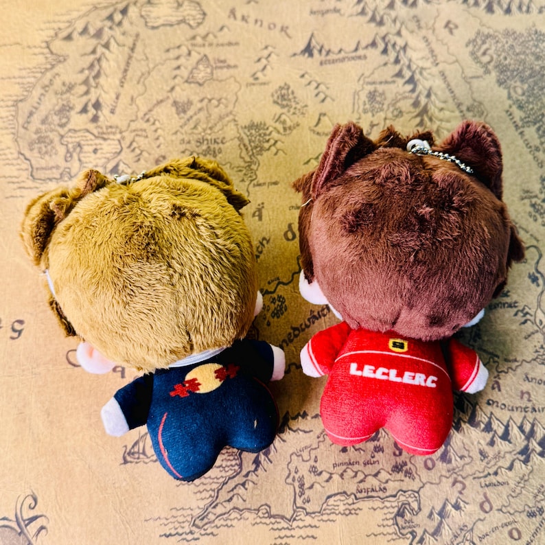 Peut inclure: Deux poup&eacute;es en peluche avec des cheveux bruns et beiges, vues de dos. L'une porte une tenue bleu marine avec un motif rouge et jaune, l'autre une tenue rouge avec "LECLERC" imprim&eacute; au dos. Les deux ont de petites cha&icirc;nes m&eacute;talliques.
