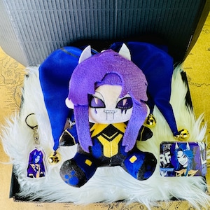 以下が含まれることがあります： 道化師の帽子、キーホルダー、小さなカードが付いたぬいぐるみ。人形は紫色の髪と青と黄色の衣装を着ています。キーホルダーとカードは同様のキャラクターデザインが特徴です。アイテムは白い毛皮の表面の黒い箱に表示されています。