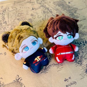 Peut inclure: Deux poup&eacute;es en peluche avec des traits de style anime. L'une a une fourrure brune et une tenue noire et marine avec le logo Red Bull. L'autre porte une combinaison de course rouge avec des accents blancs. Les deux poup&eacute;es ont de grands yeux.