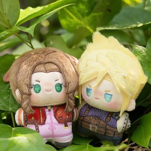 May include: Two plush dolls of anime characters. One doll has brown hair, a red jacket, and pink overalls. The other doll has spiky blonde hair, a purple top, and brown straps. Both dolls have embroidered facial features and are set against a backdrop of green leaves.