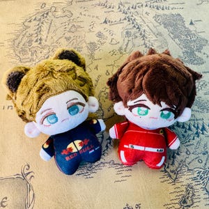 Peut inclure: Deux poup&eacute;es en peluche avec des traits de style anime. L'une a des cheveux bruns et porte une tenue bleu marine avec le texte "RED BULL". L'autre a des cheveux bruns et porte une tenue rouge avec le num&eacute;ro "16".