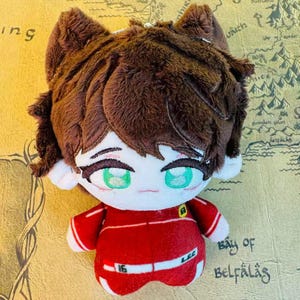 Peut inclure: Une poup&eacute;e en peluche avec un visage de style dessin anim&eacute;, des cheveux bruns et des yeux verts. La poup&eacute;e porte une tenue rouge avec des accents blancs et le num&eacute;ro "16". La poup&eacute;e a des oreilles de chat.