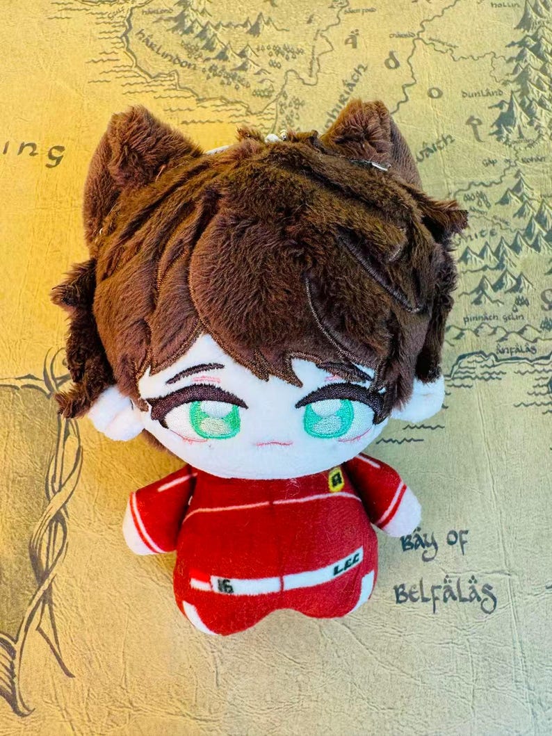 Peut inclure: Une poup&eacute;e en peluche avec un visage de style dessin anim&eacute;, des cheveux bruns et des yeux verts. La poup&eacute;e porte une tenue rouge avec des accents blancs et le num&eacute;ro "16". La poup&eacute;e a des oreilles de chat.
