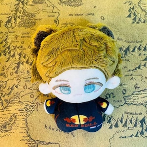 Peut inclure: Une poup&eacute;e en peluche avec une peau claire, des yeux bleus et des cheveux jaunes. La poup&eacute;e porte une tenue noire avec le logo Red Bull. Le fond est une carte avec des montagnes et du texte.