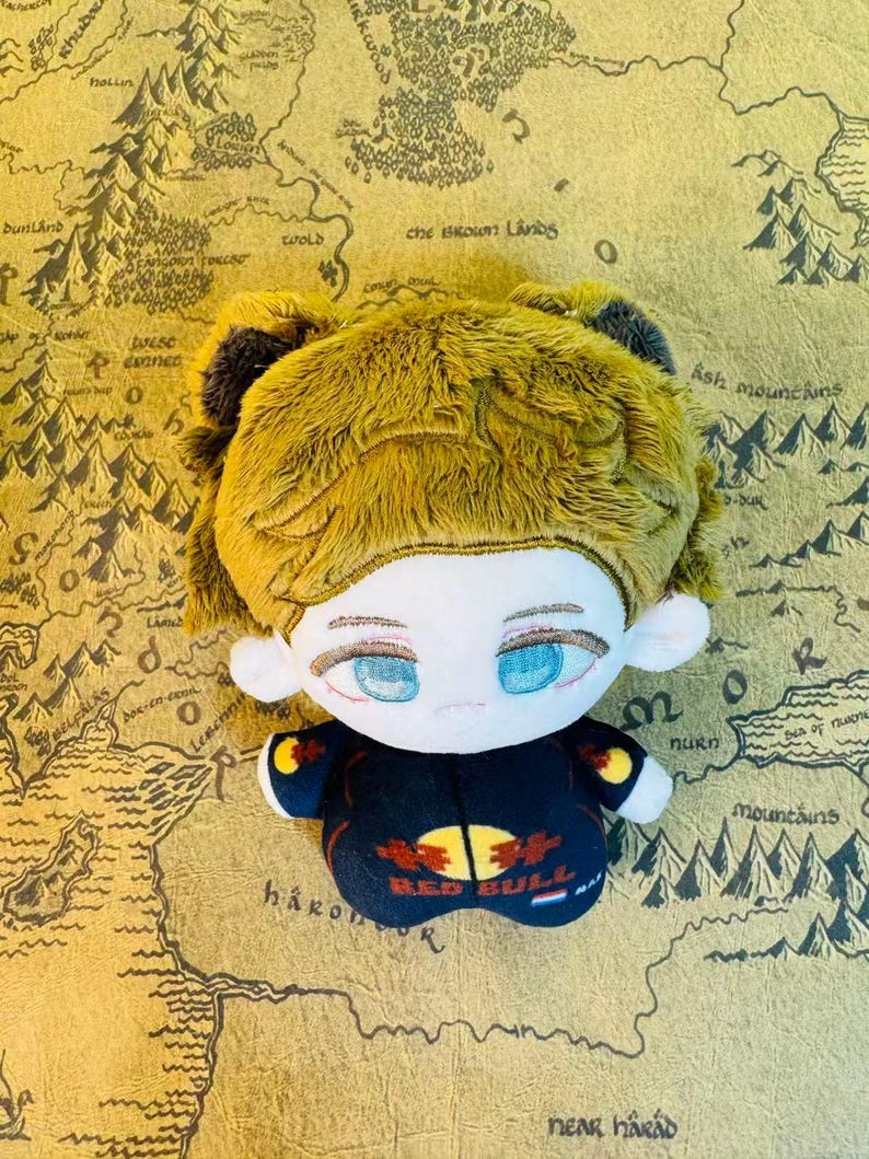 Peut inclure: Une poup&eacute;e en peluche avec une peau claire, des yeux bleus et des cheveux jaunes. La poup&eacute;e porte une tenue noire avec le logo Red Bull. Le fond est une carte avec des montagnes et du texte.