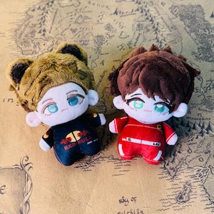 Peut inclure: Deux poup&eacute;es en peluche avec des visages brod&eacute;s et des tenues d&eacute;taill&eacute;es. L'une porte une combinaison de course Red Bull noire et rouge, l'autre un uniforme rouge avec le num&eacute;ro 16. Les deux poup&eacute;es ont les cheveux bruns et sont pos&eacute;es sur un fond de carte.