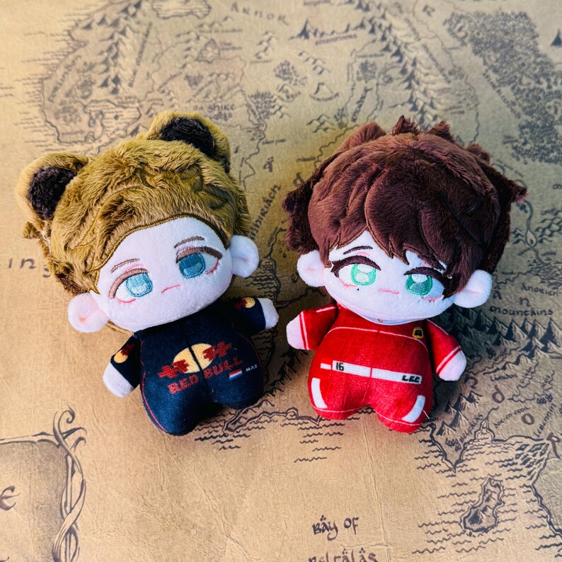 Peut inclure: Deux poup&eacute;es en peluche avec des visages brod&eacute;s et des tenues d&eacute;taill&eacute;es. L'une porte une combinaison de course Red Bull noire et rouge, l'autre un uniforme rouge avec le num&eacute;ro 16. Les deux poup&eacute;es ont les cheveux bruns et sont pos&eacute;es sur un fond de carte.