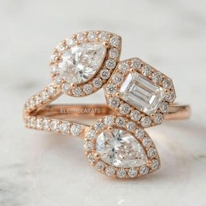 Può includere: Un anello in oro rosa con tre pietre centrali: due diamanti a forma di pera e un diamante taglio smeraldo, ciascuno circondato da un alone di piccoli diamanti rotondi. Anche la fascia è incastonata con piccoli diamanti rotondi. Il testo ELEUNECARATS è visibile.