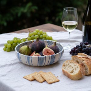 Puede incluir: Un cuenco de cerámica con rayas azules contiene higos frescos, algunos cortados para revelar su interior rojo. Uvas verdes, galletas y pan rebanado están dispuestos sobre un mantel blanco, junto con una copa de vino blanco y una botella.