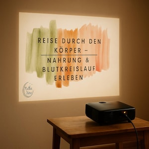 May include: A black projector on a wooden table projects text and watercolor brushstrokes onto a wall. The projected image includes the text "REISE DURCH DEN KÖRPER - NAHRUNG & BLUTKREISLAUF ERLEBEN" and a logo.