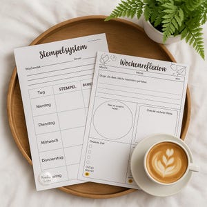 Könnte beinhalten: Zwei weiße Papierbögen mit deutschem Text, einer mit der Aufschrift "Stempelsystem" und der andere mit "Wochenreflexion", liegen auf einem Holztablett. Eine Tasse Kaffee mit Latte Art steht auf einer Untertasse. Eine kleine Topfpflanze befindet sich im Hintergrund.