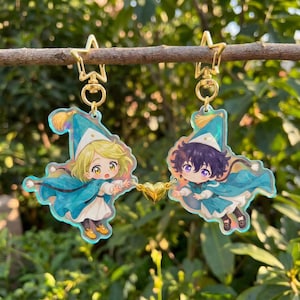 Könnte beinhalten: Zwei Schlüsselanhänger aus Acryl mit Anime-Charakteren. Jeder Anhänger hat einen goldfarbenen sternförmigen Verschluss. Die Charaktere tragen blaue Umhänge und Hüte mit blonden und lila Haaren. Ein goldenes Herz verbindet die beiden Anhänger.