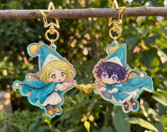 May include: Two acrylic charm keychains featuring anime-style characters. Each charm has a gold-colored star-shaped clasp. The characters are wearing blue capes and hats, with blonde and purple hair. A gold heart connects the two charms.