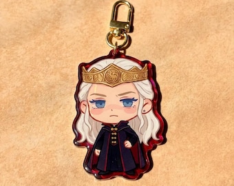 May include: An acrylic keychain featuring a stylized illustration of a character with long white hair, blue eyes, and a gold crown. The character wears a dark cloak with red accents. The charm is attached to a gold-colored metal lobster clasp.