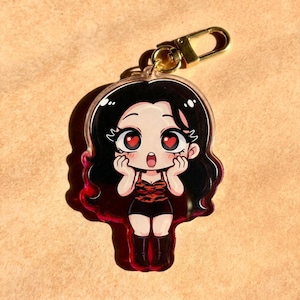May include: An acrylic keyring featuring a cartoon character with long black hair, large eyes with heart-shaped pupils, and a surprised expression. The character wears a red and black tiger-print top, black shorts, and black boots, attached to a gold clasp.
