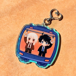 May include: A clear acrylic keychain shaped like a train car window, featuring an illustration of two anime-style characters standing inside a train. The characters are holding onto overhead hand straps. It includes a silver heart-shaped clasp.