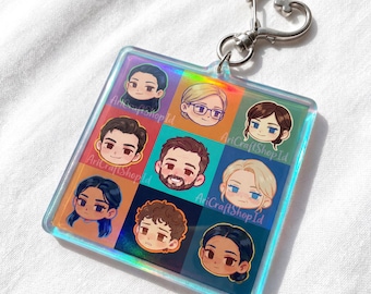 May include: Square keychain with a heart-shaped clasp. The keychain features a grid of nine cartoon portraits with different hair colors and styles. Each portrait is set against a different colored background. The text "AriCraftShopId" is visible on the keychain.