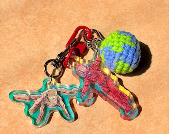 May include: A collection of keychains featuring space-themed designs. One keychain depicts an astronaut in a red spacesuit, another shows a rock formation, and a third is a crocheted blue and green globe. The keychains are attached to a red carabiner.