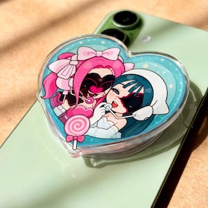 Puede incluir: Un soporte para teléfono en forma de corazón con una colorida ilustración estilo anime de dos personajes con cabello rosa y verde azulado, con lazos y auriculares, compartiendo una paleta rosa sobre un fondo azul claro con estrellas.