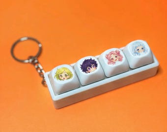 May include: A white keychain featuring four keycaps with anime-style character illustrations. The keycaps are arranged in a row and attached to a silver keyring. The background is a solid orange color.