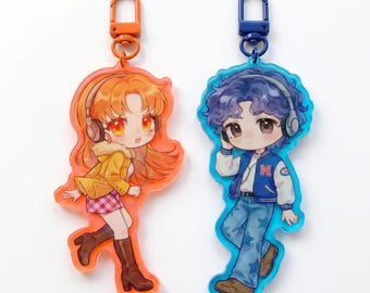 May include: Two acrylic keychains featuring anime-style characters with headphones. The left keychain is orange-tinted with a character in a yellow jacket and plaid skirt. The right keychain is blue-tinted with a character in a blue varsity jacket.
