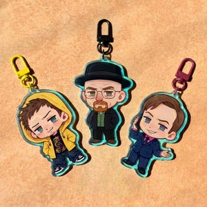 Op de afbeelding: Drie acryl sleutelhangers met stripfiguur-achtige personages. Eén toont een persoon in een gele hoodie, één een persoon met een zwarte hoed en jas, en één een persoon in een blauw pak. Elke hanger heeft een gekleurde plastic sluiting in geel, zwart of paars.