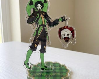 May include: An acrylic standee featuring a jester character in a green and black outfit with a matching hat, holding a small dangling charm of a white-haired figure in a red hat. The standee is mounted on a circular green plaid base.