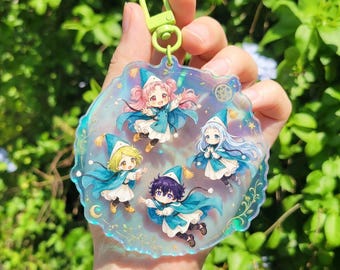May include: A clear, iridescent keychain featuring five cartoon characters in blue and white outfits with matching hats. The keychain has a gold-colored clasp and is held against a backdrop of green foliage.