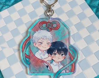 May include: An acrylic charm featuring a chibi-style illustration of two characters with white and black hair, one poking the other's cheek. The charm has a holographic finish, a red decorative knot design at the top, and a silver heart-shaped clasp.