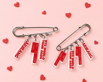 May include: Two silver-toned metal safety pins, each featuring three dangling rectangular acrylic charms. The charms are printed with red text reading ARIRANG, J-HOPE, JIN, SUGA, and BTS WORLD TOUR 2026. The pins are arranged against a light pink background.