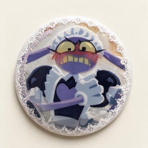 Pode incluir: Um broche circular com um personagem roxo em uma roupa de empregada com gola e avental brancos com babados. O personagem tem uma expressão de raiva com bochechas coradas. O texto ©2023 The Amazing Digital Circus está impresso na borda esquerda e o broche é contornado por uma renda branca.