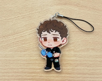 May include: A clear acrylic charm featuring a cartoon doctor with brown curly hair, wearing a black uniform, blue gloves, and holding a scalpel. The charm has a silver-colored clasp and a black lanyard.