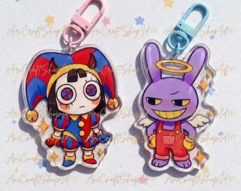 May include: Two acrylic keychains featuring cartoon characters. One is a jester in red, blue, and yellow with a pink clasp. The other is a purple bunny with a halo and wings, wearing red overalls, with a blue clasp.