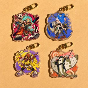 May include: Four acrylic keychains featuring colourful cartoon characters from Splatoon. The top left shows a character with teal hair and a weapon, the top right a blue-haired character with a fan, the bottom left a yellow-haired character, and the bottom right a manta ray creature.
