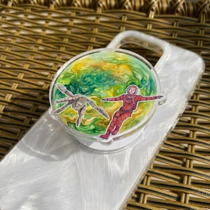 May include: A clear acrylic phone grip featuring a colorful illustration of an astronaut in a red spacesuit and a creature against a green and yellow planet background. The grip is attached to a white phone case.