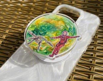 May include: A clear acrylic phone grip featuring a colorful illustration of an astronaut in a red spacesuit and a creature against a green and yellow planet background. The grip is attached to a white phone case.