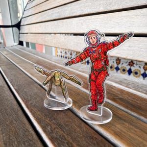 May include: Two clear acrylic standees featuring illustrated characters. One is an astronaut in a bright red spacesuit with a helmet, and the other is a small, tan, multi-limbed creature with green accents. Both figures are mounted on circular bases.