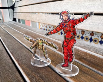 May include: Two clear acrylic standees featuring illustrated characters. One is an astronaut in a bright red spacesuit with a helmet, and the other is a small, tan, multi-limbed creature with green accents. Both figures are mounted on circular bases.