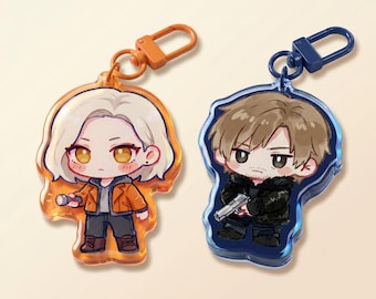 May include: Two acrylic keychains featuring cartoon characters. One keychain has a character with blonde hair, wearing an orange jacket, and holding a small object. The other keychain has a character with brown hair, wearing a black jacket, and holding a gun. Both have colored clasps.