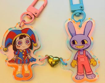 May include: Two cartoon character keychains. One keychain features a jester character in red, blue, and yellow. The other keychain features a purple rabbit character in pink overalls and yellow gloves. Both keychains are connected by a gold heart charm.