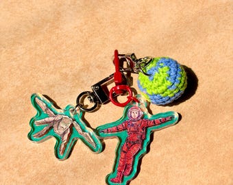 May include: Keychains with space-themed designs. One keychain features a red astronaut, another a green alien-like shape, and a third a blue and green crocheted globe. All keychains are attached to a silver clasp with a red accent.