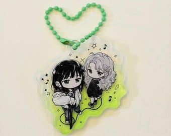 May include: A clear acrylic keychain featuring two chibi-style characters in black and white, one with straight dark hair and one with wavy light hair, set against a lime green background with musical notes and stars, attached to a green ball chain.
