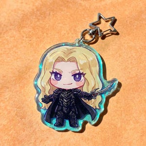 Puede incluir: Un llavero de acrílico con un personaje estilo chibi de cabello rubio largo, ojos morados y armadura negra, sosteniendo una pequeña espada plateada. El personaje está impreso en una pieza de acrílico transparente con borde iridiscente.