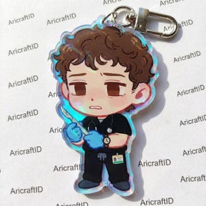 May include: An acrylic keyring featuring a chibi-style character with brown curly hair, wearing black scrubs, a stethoscope, and blue gloves. The character holds a scalpel and has an ID badge attached to the waist. The charm has a holographic finish.