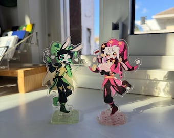 May include: Two acrylic standee figures featuring stylized characters in jester-inspired outfits. The left figure wears green and black with rabbit ears, while the right figure wears pink and black with long ears, both standing on clear bases.