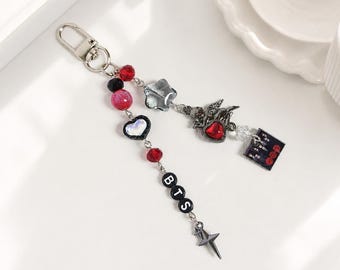 May include: A silver-toned keychain featuring a metal clasp, red and black beads, a heart-shaped charm, a star-shaped bead, a metallic cherub charm with a red heart, a square charm with a graphic print, and black letter beads spelling BTS.