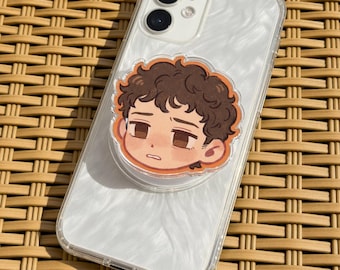 May include: A clear phone case with a white pearlescent design, featuring a pop-up grip with a cartoon illustration of a person's face. The phone case is on a woven surface.